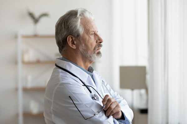 Thoughtful serious senior doctor looking through window lost in thoughts. Worried pensive old physician thinking of healthcare question, concerned of challenge, feels anxious makes difficult decision.